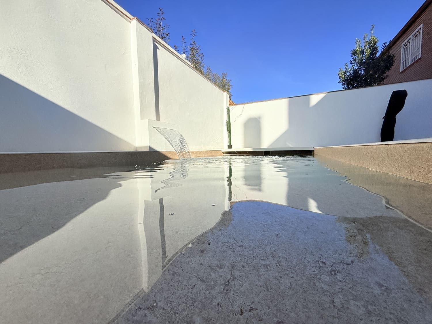 Fuente con paredes blancas, borde de piedra y cielo azul. El agua fluye hacia una piscina poco profunda.