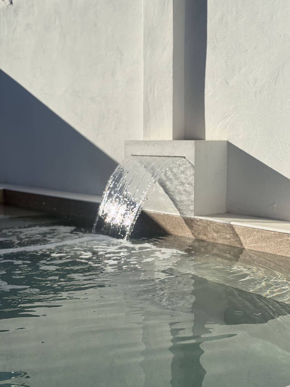 Agua cayendo en cascada desde un caño blanco hacia una piscina. Paredes blancas, molduras color canela claro.