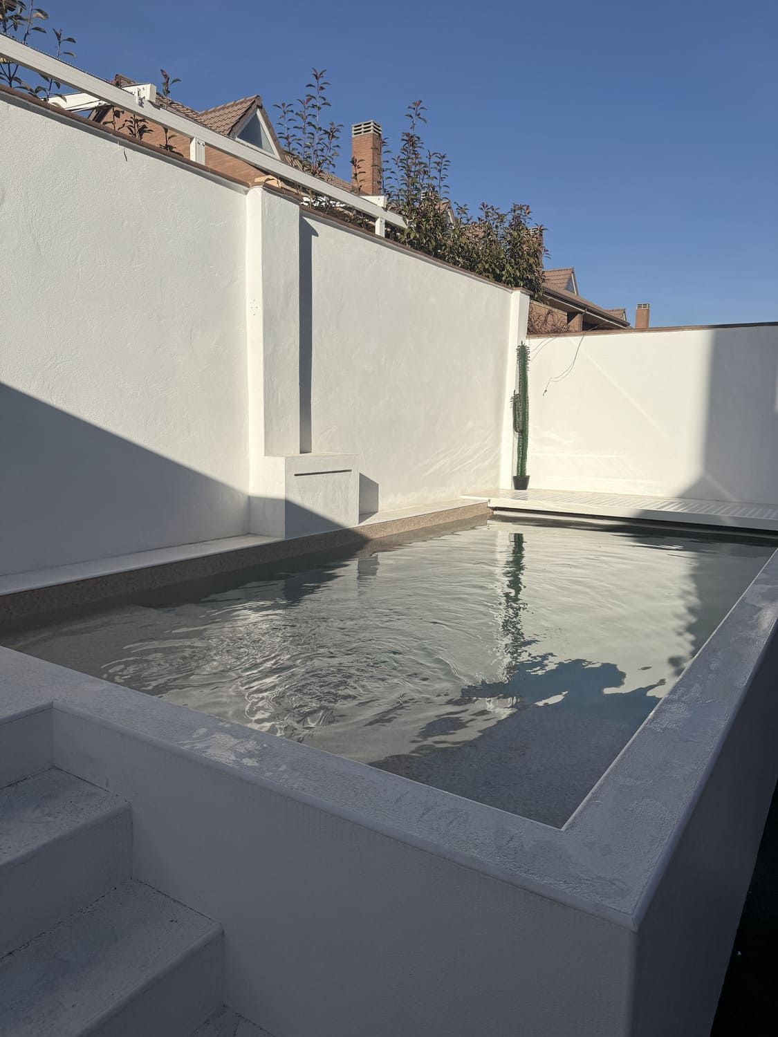 Pequeña piscina blanca y rectangular con escalones en un patio. Paredes blancas y cielo azul.
