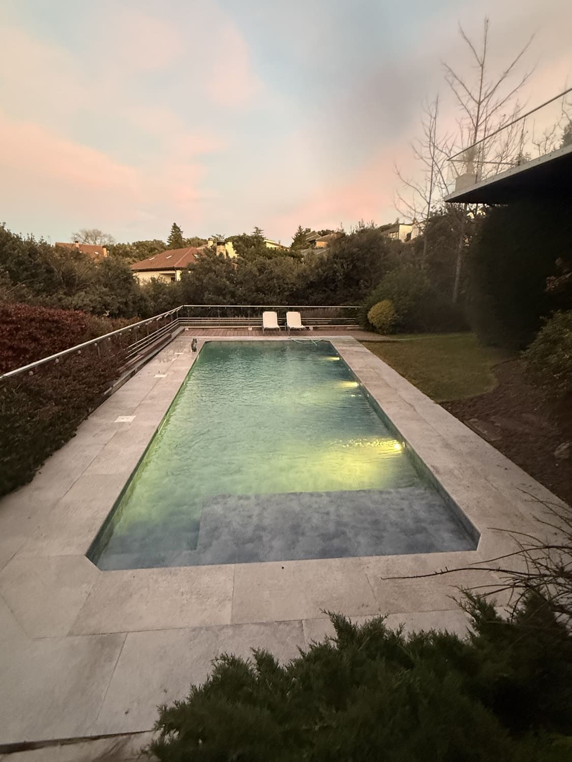 Piscina rectangular al atardecer, rodeada de un patio de piedra, una exuberante vegetación y un cielo teñido de rosa.