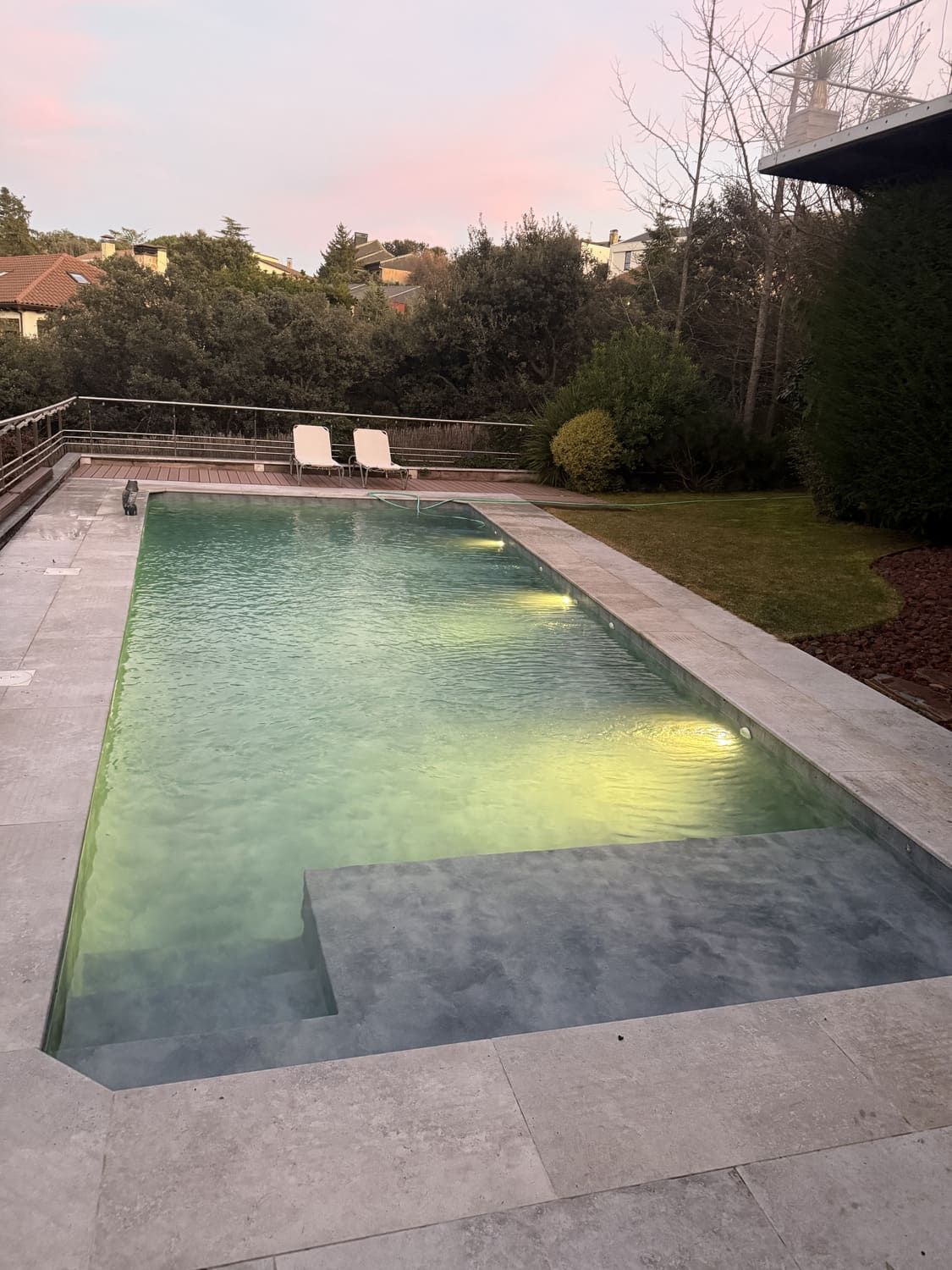 Piscina rectangular al atardecer, con escalera de obra, rodeada de patio de piedra y jardinería.