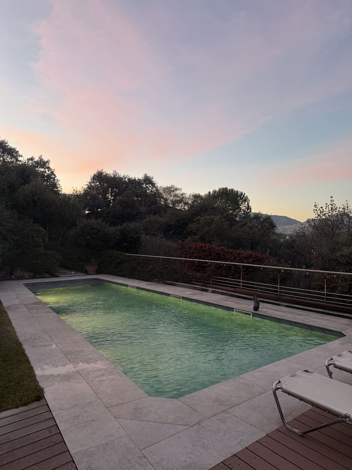 Piscina al atardecer con cielo colorido, rodeada de árboles y un deck de madera.