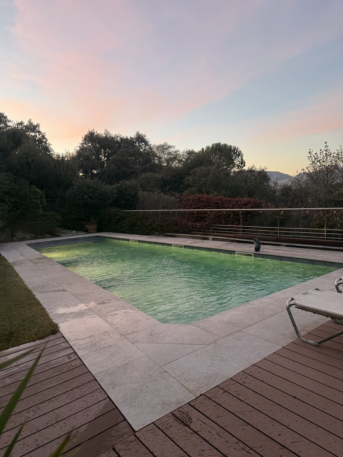 Piscina con terraza de madera, exuberante vegetación y cielo al atardecer.