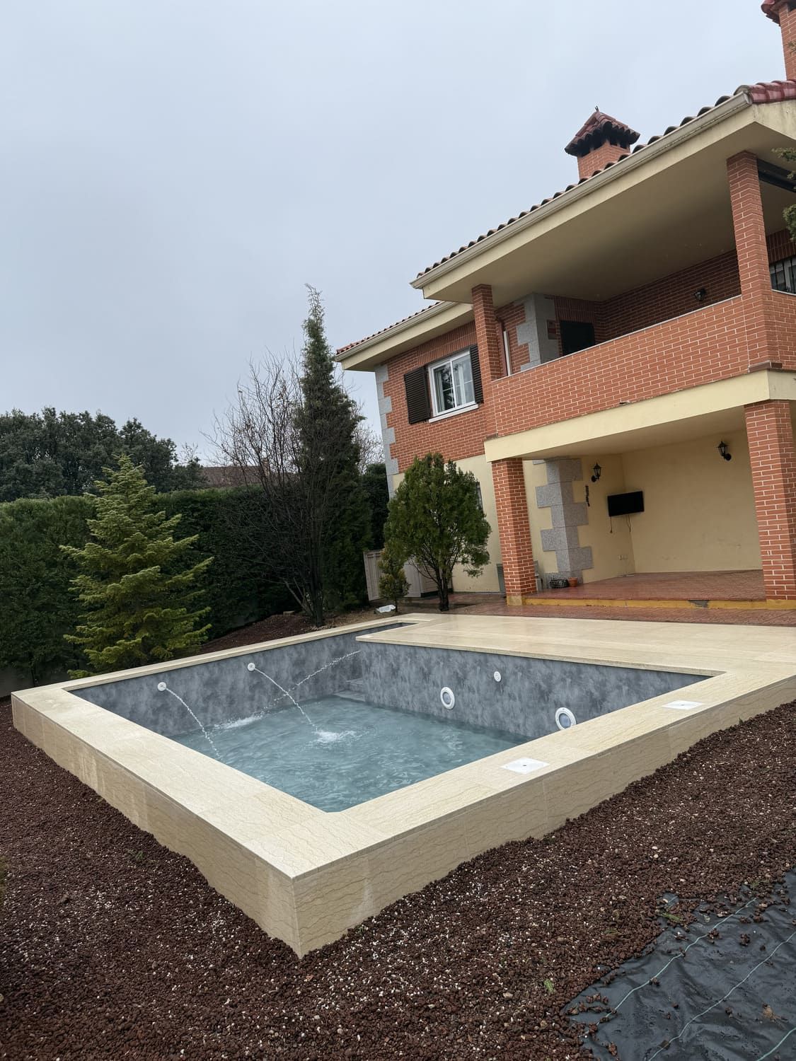 Piscina cuadrada junto a una casa de ladrillo en un día nublado. Piscina oscura con borde de hormigón color canela y mantillo marrón.