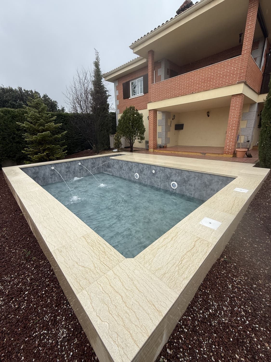 Piscina rectangular vacía con borde de piedra, frente a una casa de ladrillo de dos pisos con balcón, cielo nublado.