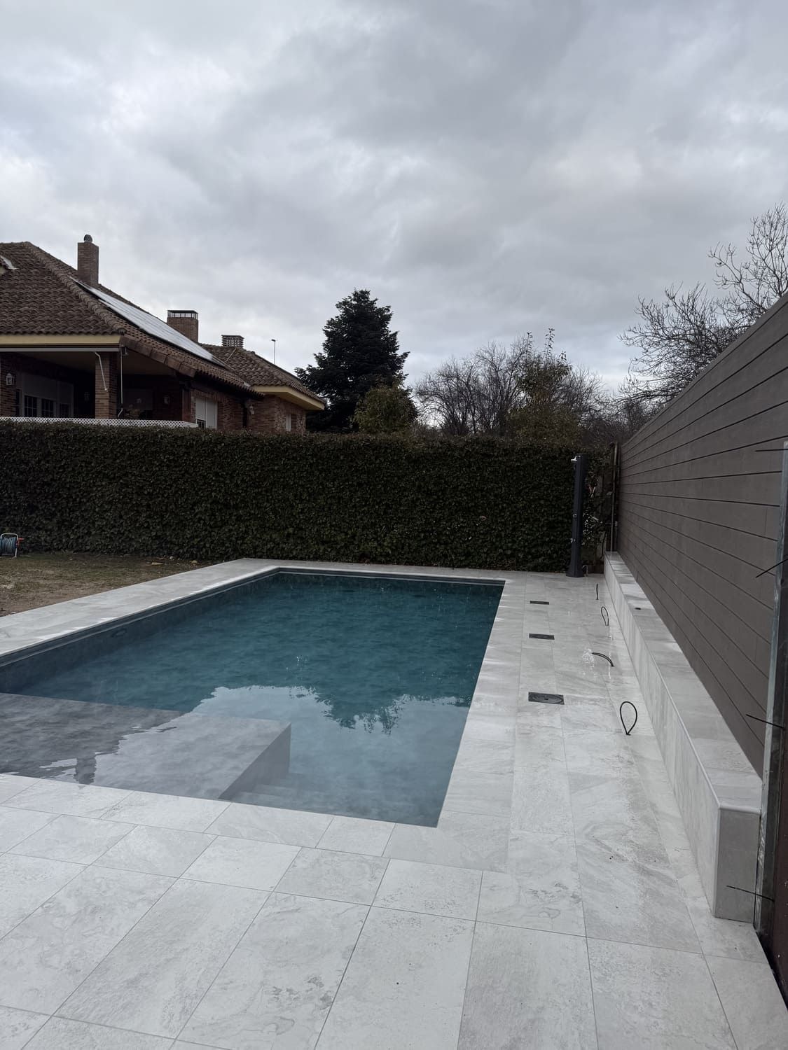 Piscina exterior con azulejos gris claro, agua oscura y cielo nublado. Seto verde y valla de madera al fondo.