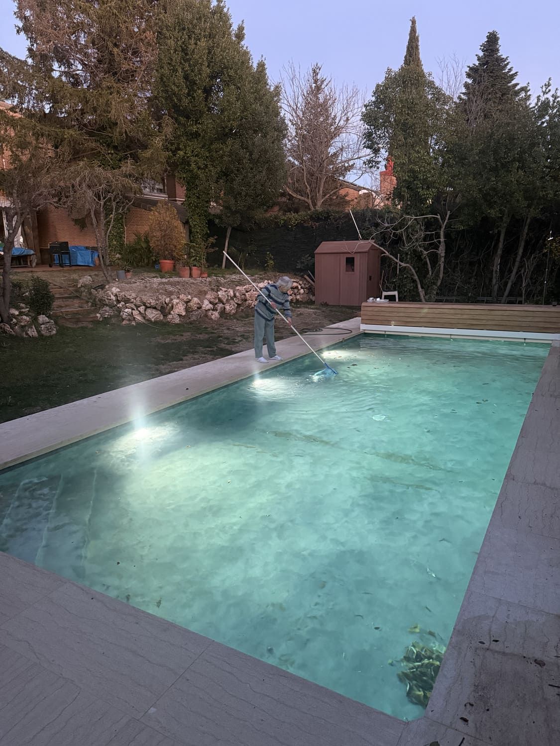 Persona limpiando una piscina rectangular al aire libre con un cepillo de mango largo.