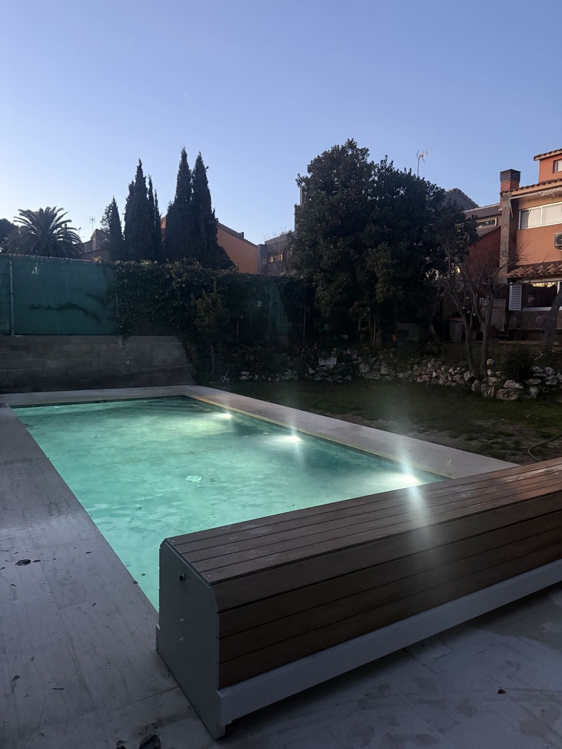 Piscina con banco revestido de madera, iluminada con luces sumergidas. Árboles y casas al fondo.