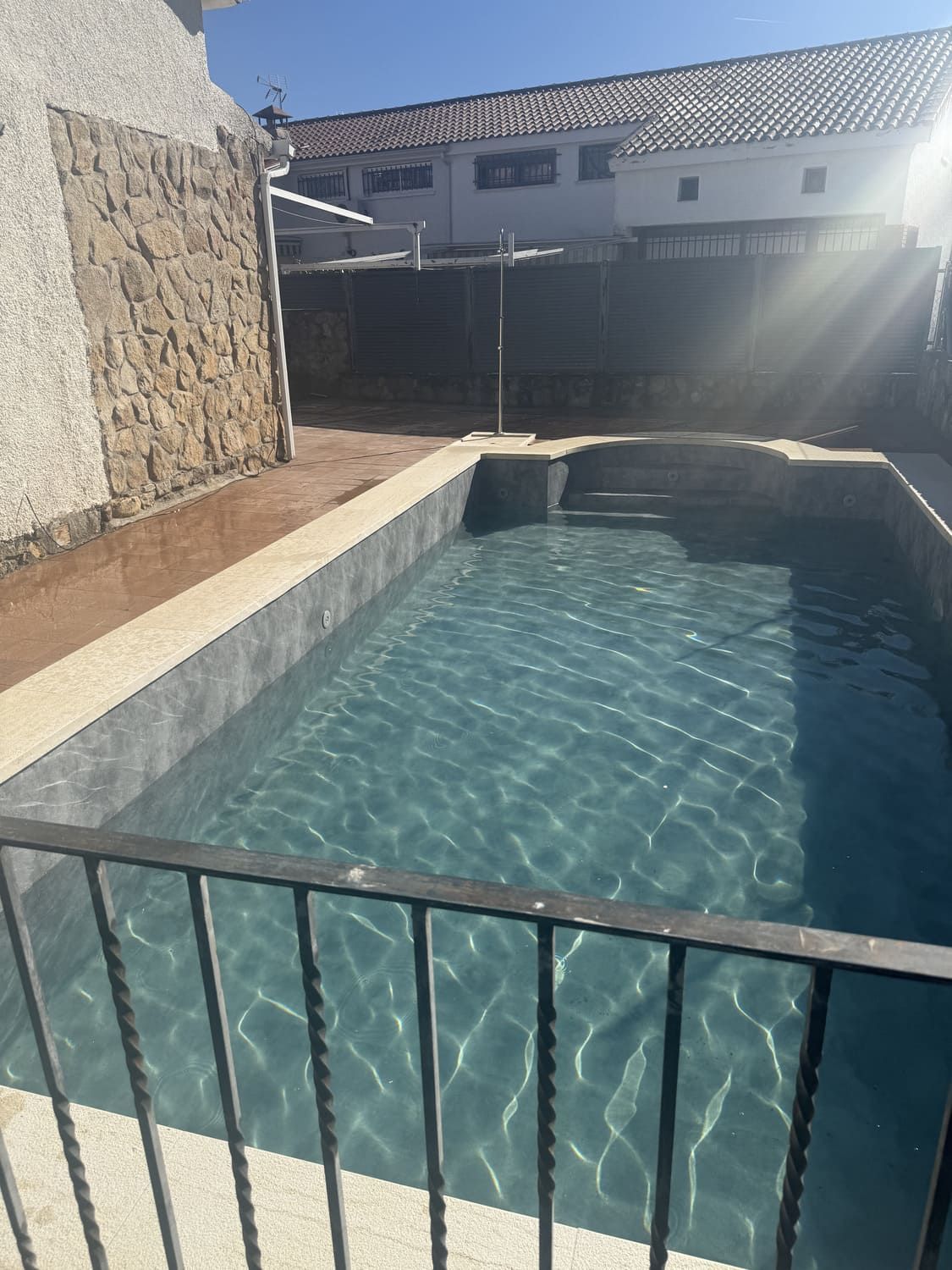 Piscina rectangular de agua cristalina, rodeada de un borde de hormigón y una valla metálica negra, bajo un cielo luminoso.