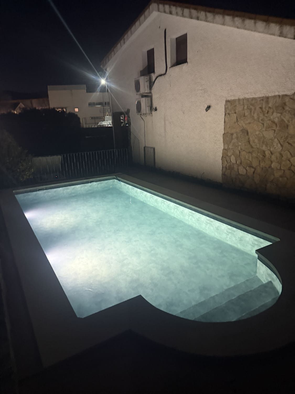 Una vista nocturna de una piscina iluminada junto a un edificio blanco.