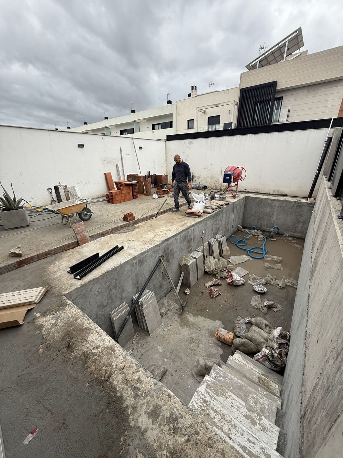 Construcción de una piscina en una azotea. Un trabajador se encuentra dentro de la piscina inacabada, rodeado de materiales. Cielo nublado.