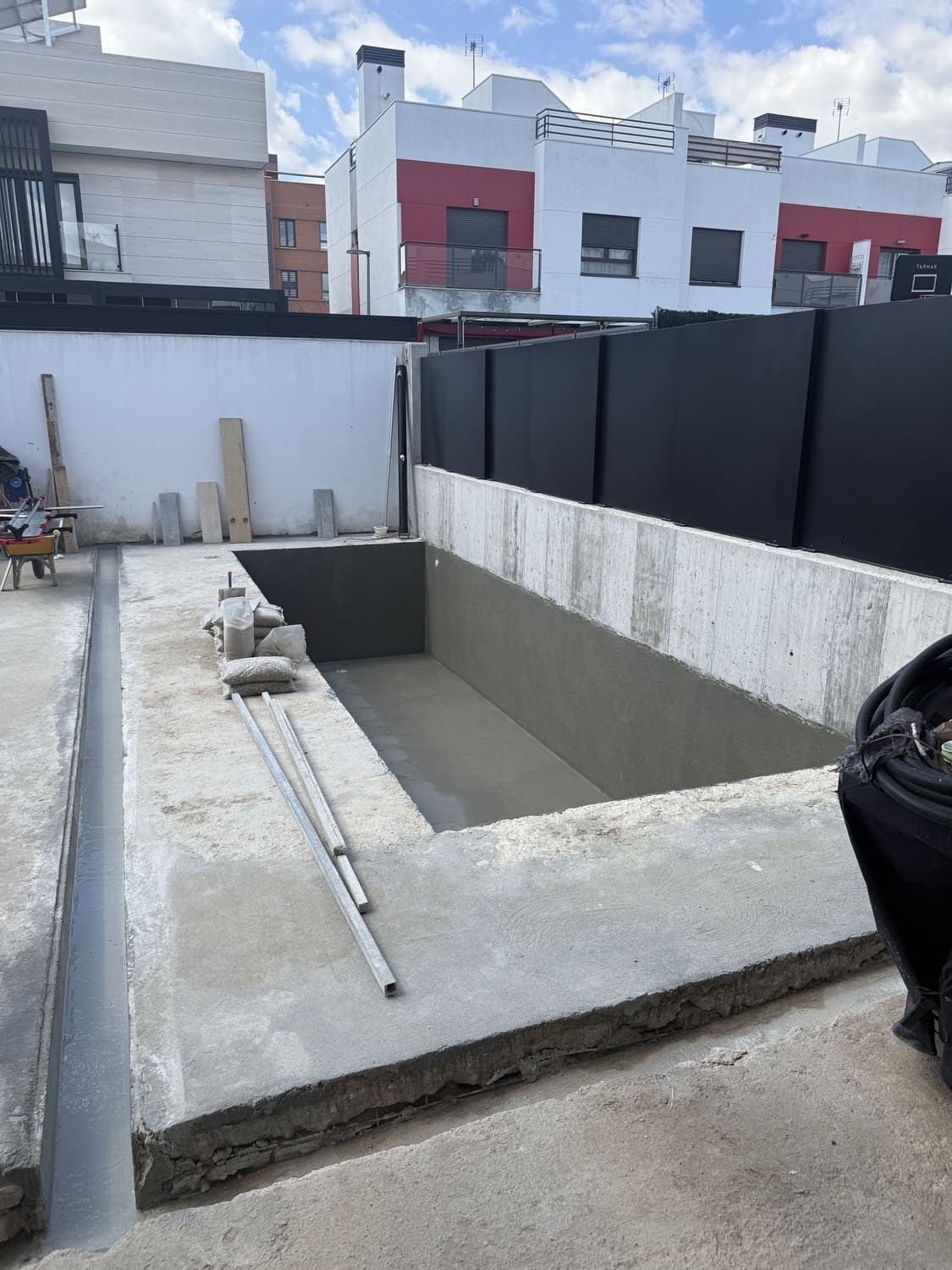 Piscina de concreto en construcción, paredes de cemento gris, tuberías, reja negra al fondo, casas adosadas.