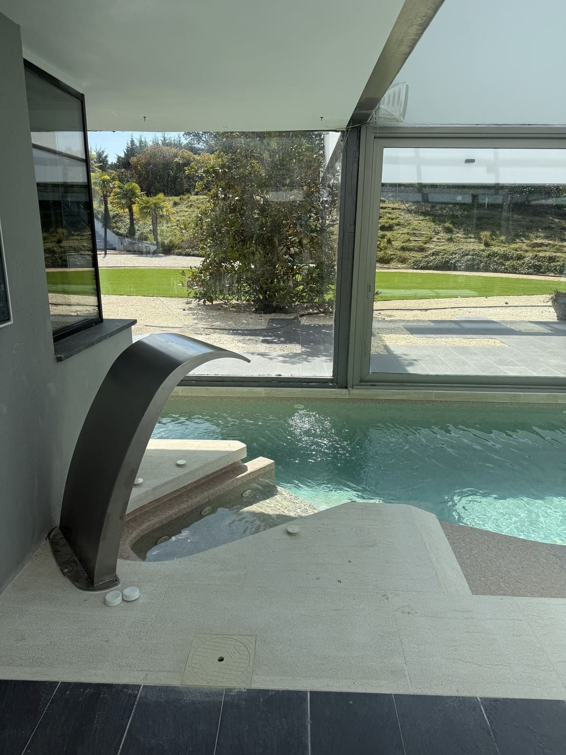 Una piscina moderna con cascada, escalones y grandes ventanales con vistas a la vegetación.