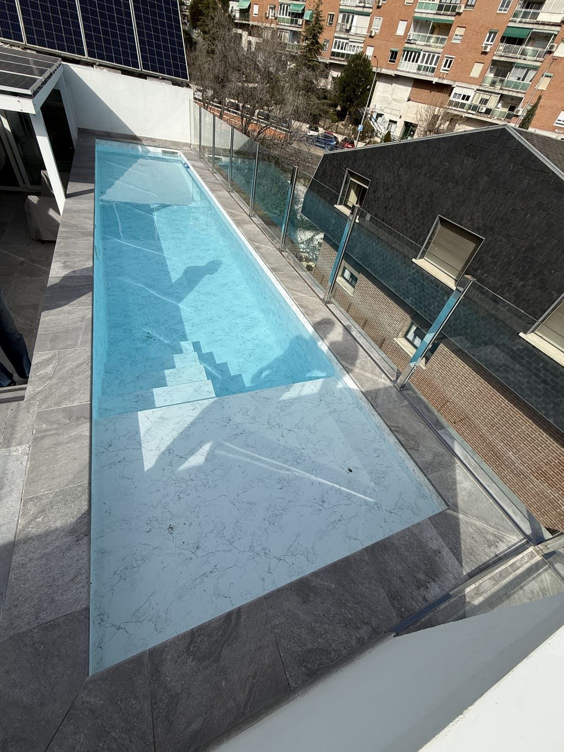 Piscina en la azotea con paredes de cristal transparente, llena de agua azul. Patio de piedra con edificios de la ciudad al fondo.