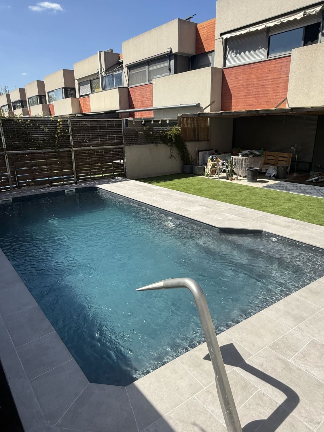 Piscina rectangular en un patio trasero con casas adosadas al fondo. Agua azul, azulejos grises, césped verde. Día soleado.