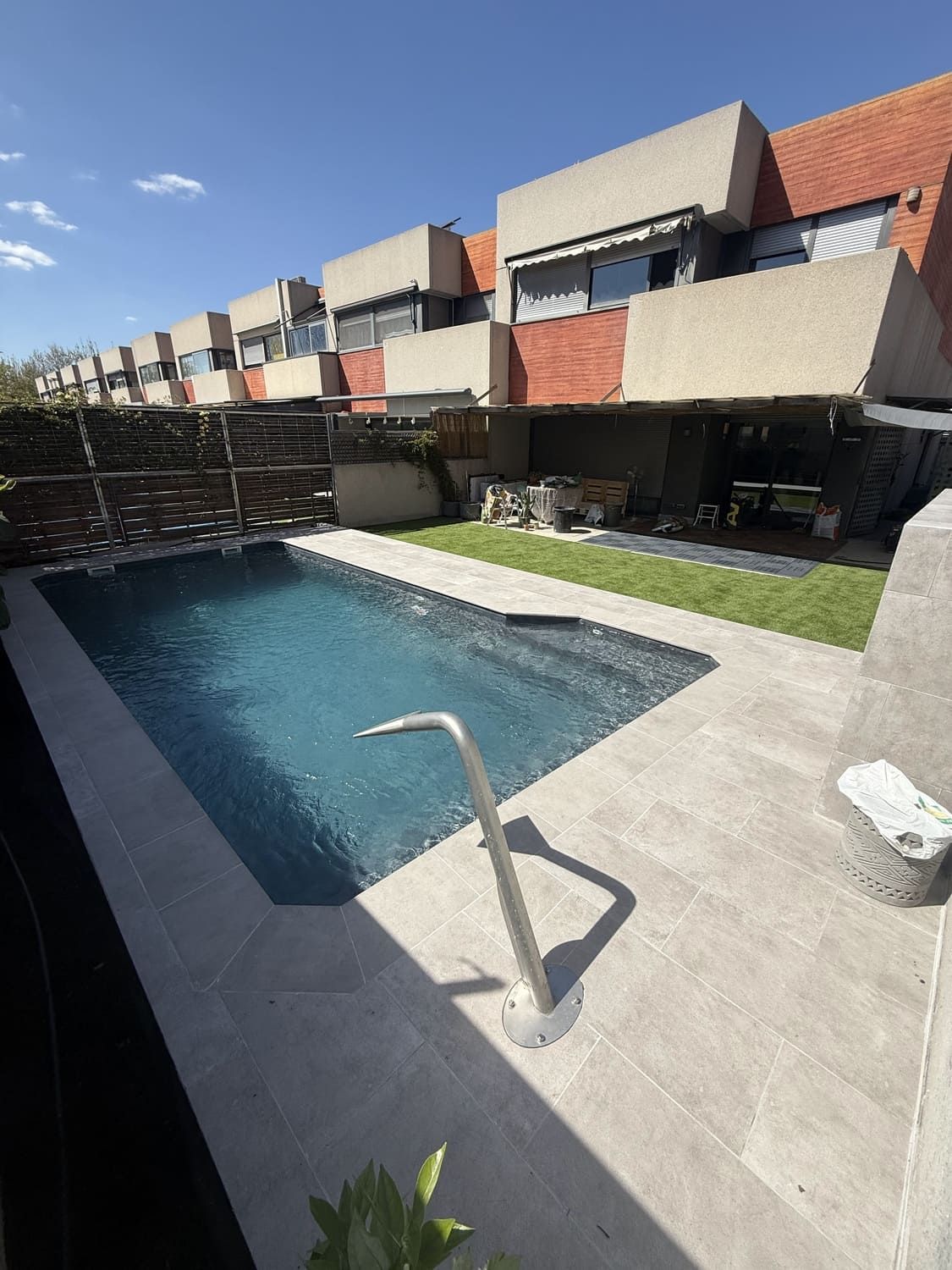 Pequeña piscina en un patio trasero con edificios al fondo y un cielo soleado.