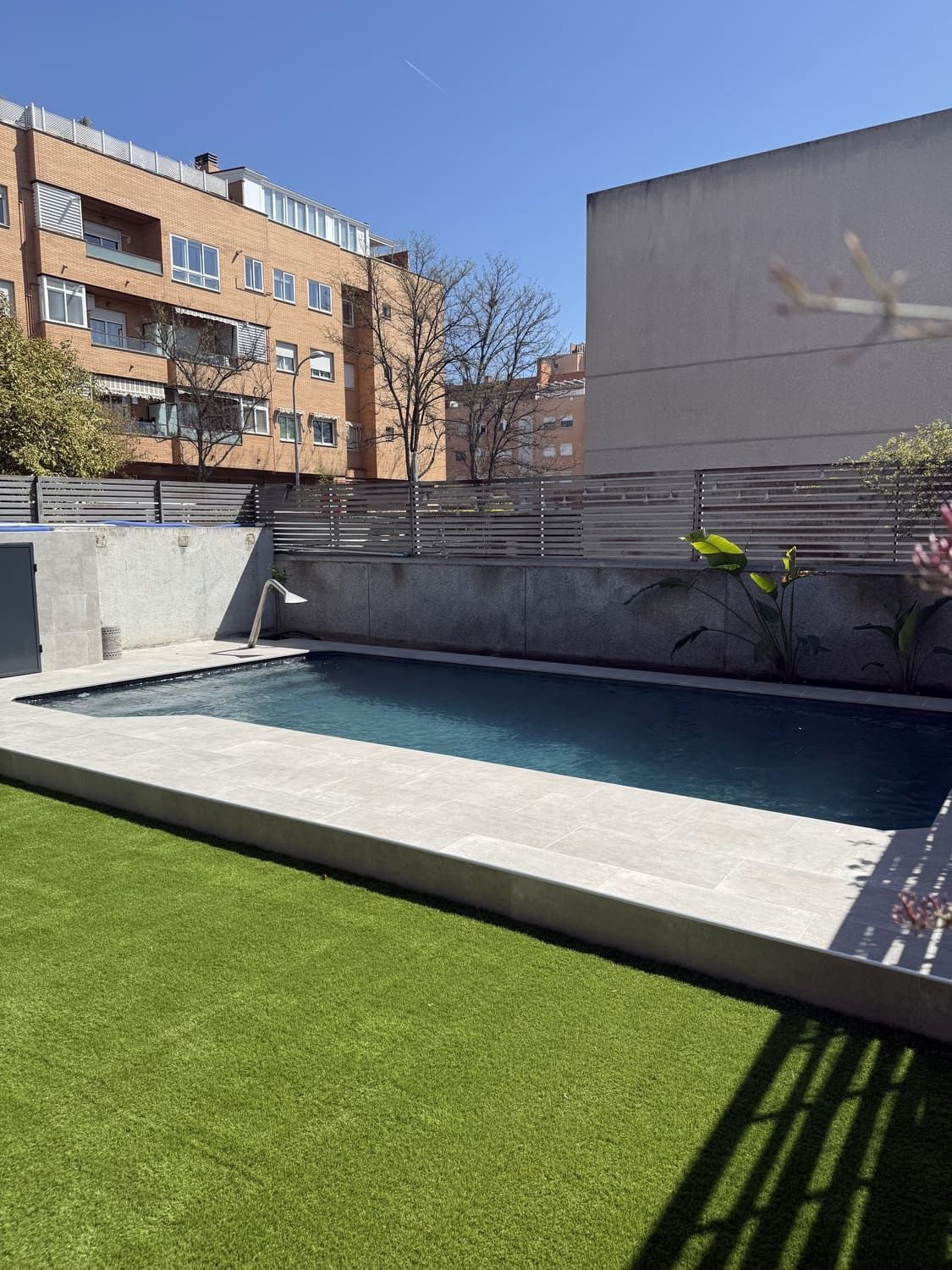 Patio trasero con piscina rectangular, rodeado de césped verde y concreto, con edificios de departamentos al fondo en un día soleado.