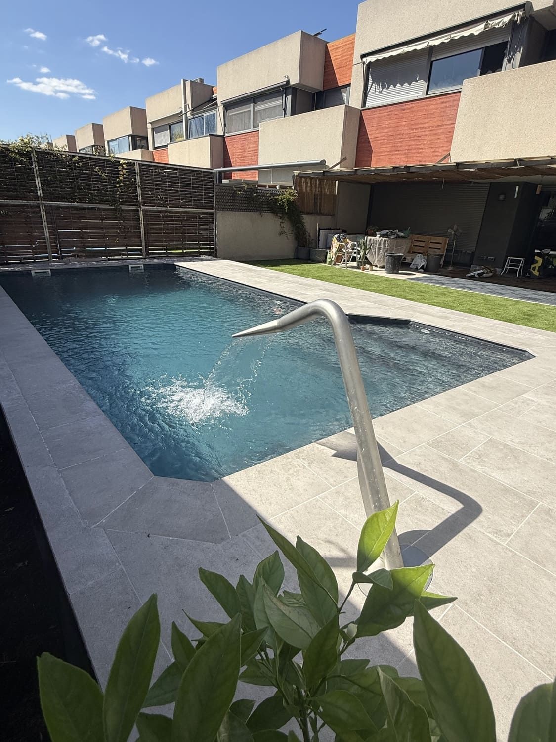 Piscina con fuente en un patio residencial. Edificios con ventanas al fondo. Día soleado.