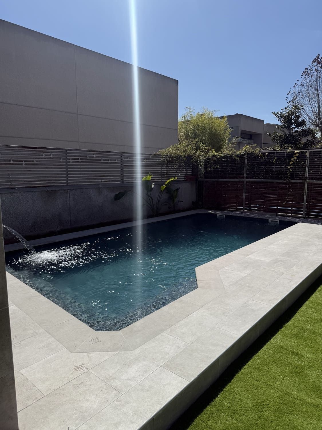 Piscina con superficie azul oscuro, rodeada de hormigón y césped verde bajo un cielo brillante y soleado.