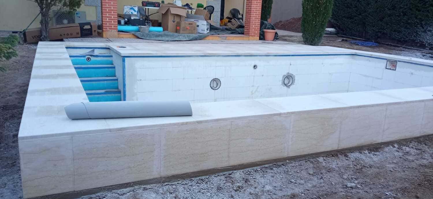 Piscina vacía en construcción. Borde de piedra beige y escalones azules. Materiales de construcción visibles al fondo.