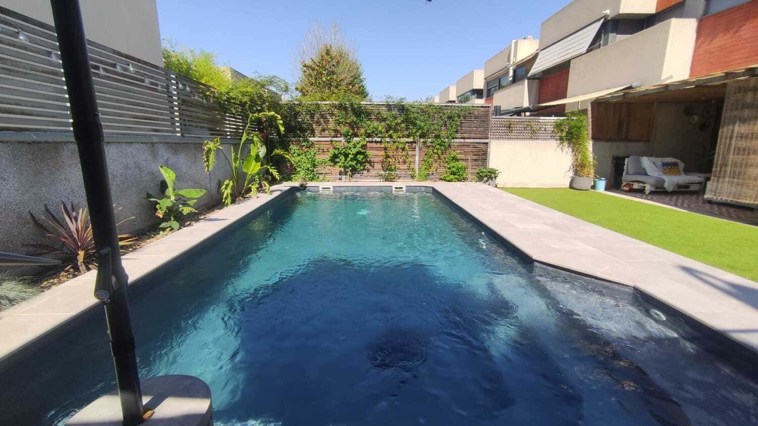 Piscina rectangular en el patio trasero con césped verde, arbustos y casas al fondo.