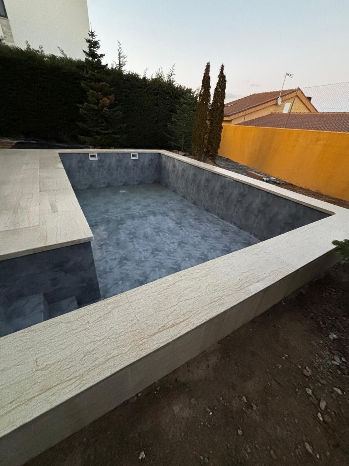 Piscina rectangular vacía con borde de piedra clara e interior gris oscuro. Resaltada por arbustos verdes y una pared amarilla.
