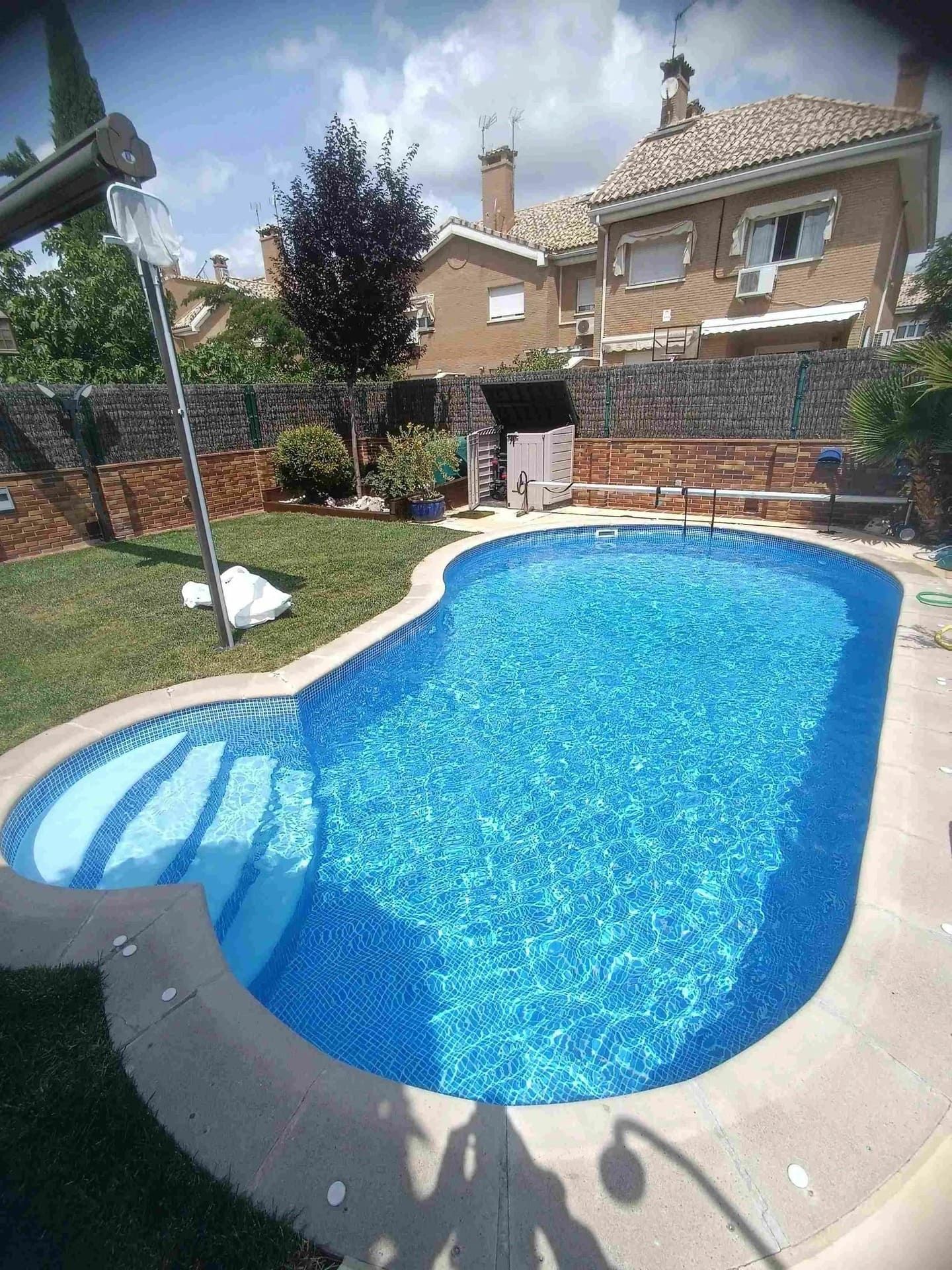 Piscina con gresite azul, rodeada de césped y con un edificio de ladrillo al fondo.