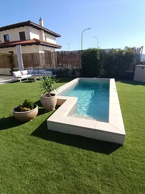 Patio trasero con piscina rectangular; molduras de piedra beige; césped verde; casa beige al fondo.