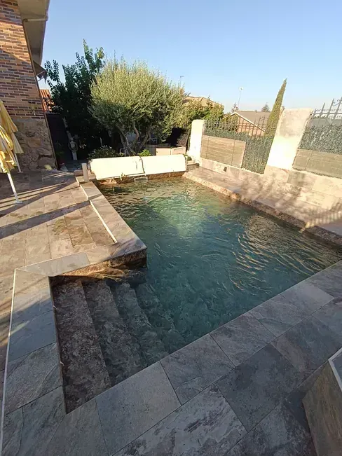 Piscina rectangular con escalones de piedra. El agua cristalina refleja la luz del sol. Edificio de ladrillo al fondo.