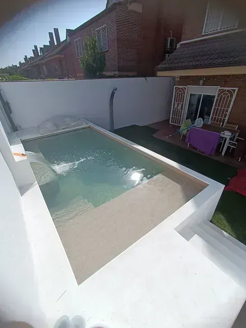 Una piscina rectangular en un patio de paredes blancas. Se ve una casa al fondo. Día soleado.