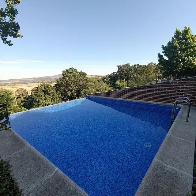 Piscina con vistas a un paisaje con árboles y una estructura arqueada lejana bajo un cielo azul claro.