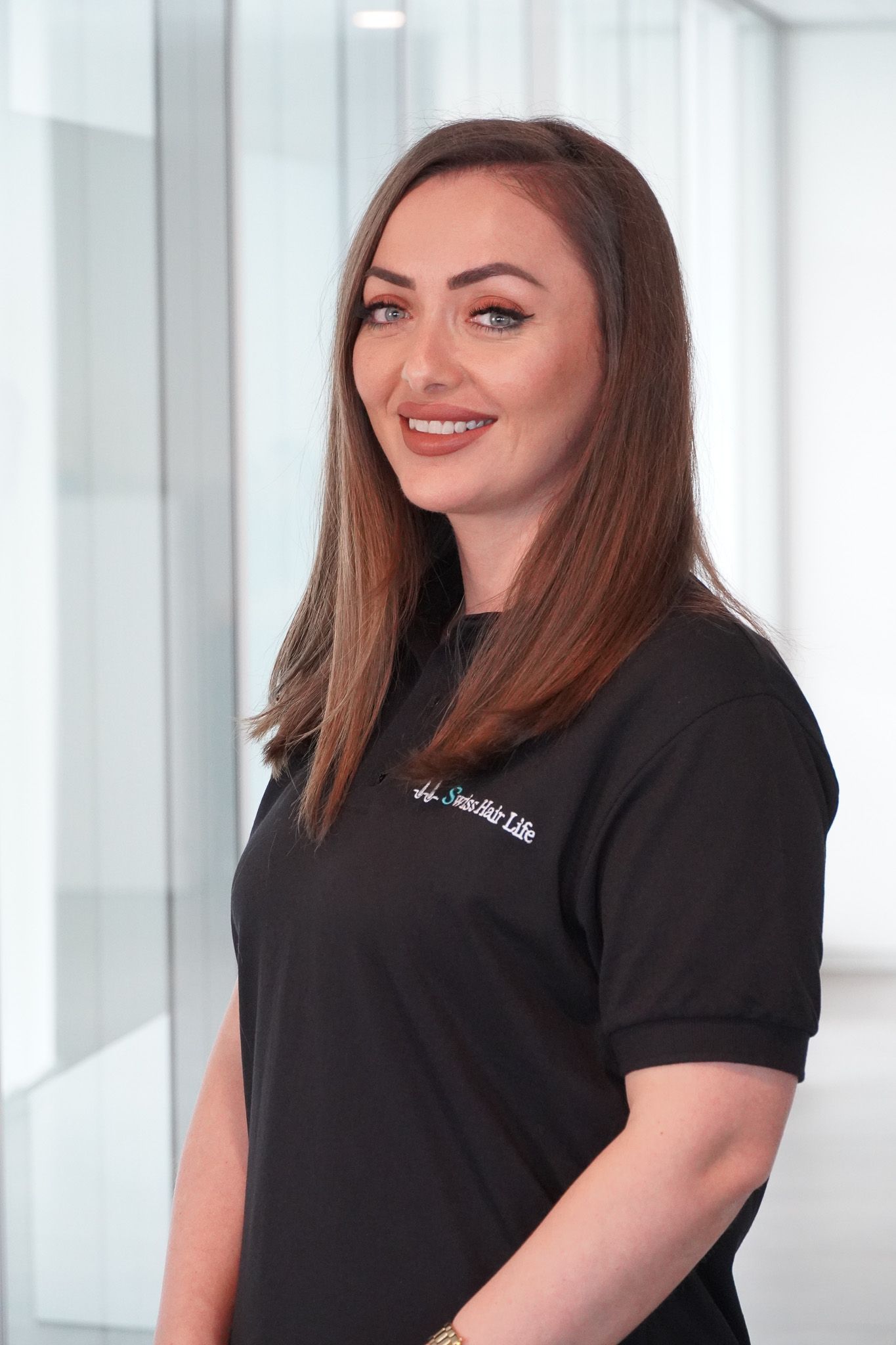 Frau mit langen braunen Haaren lächelt, trägt ein schwarzes Hemd und steht in einem Büro.