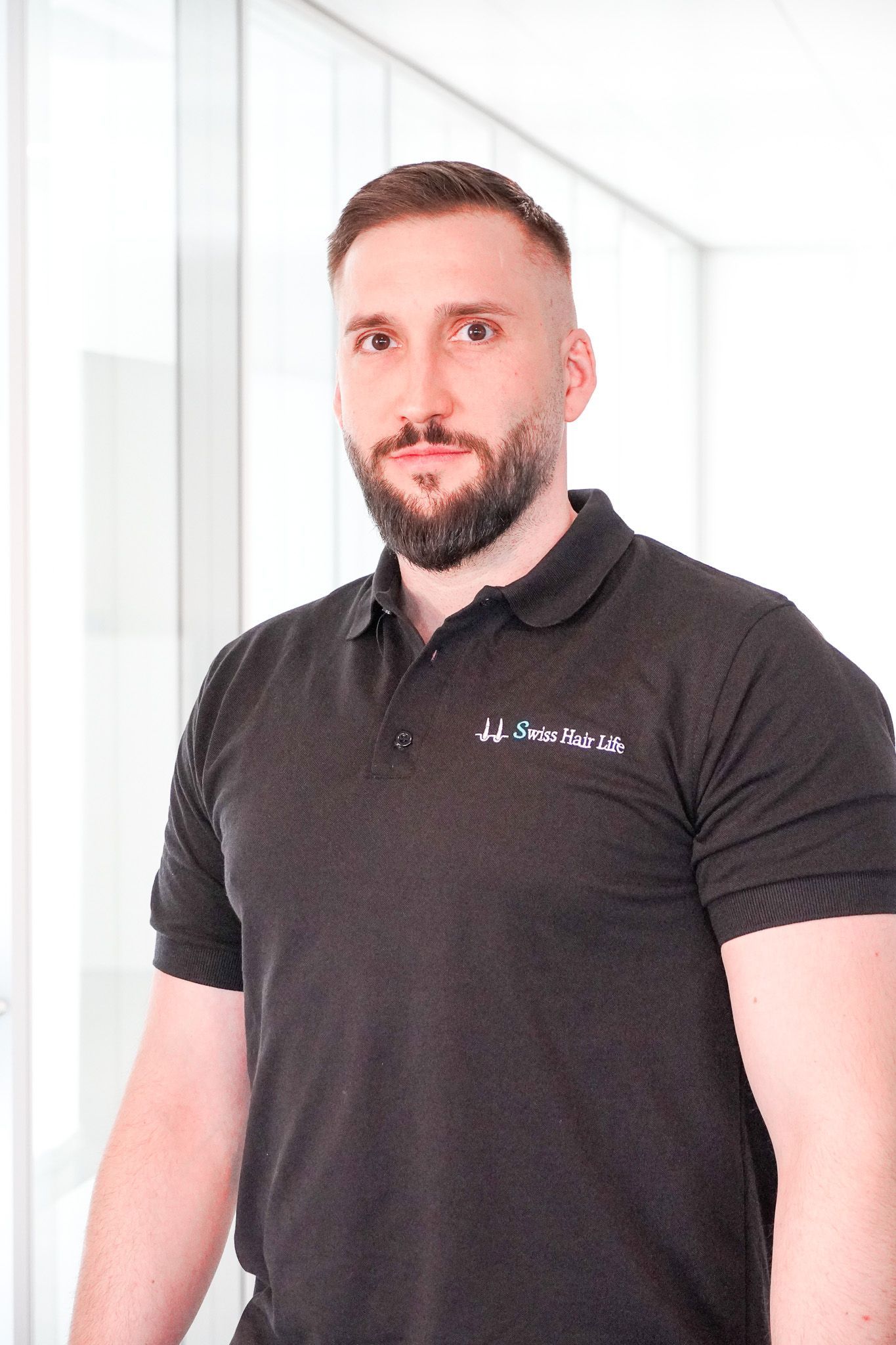 Mann mit Bart und schwarzem Poloshirt steht drinnen in der Nähe eines Fensters.