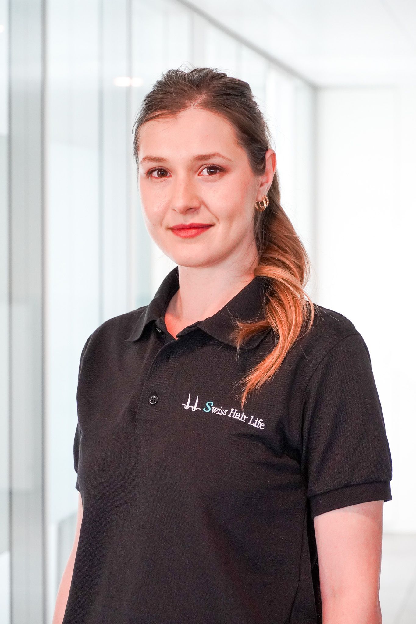 Frau mit braunem Haar in einem schwarzen Poloshirt, lächelnd in einem modernen weißen Flur.