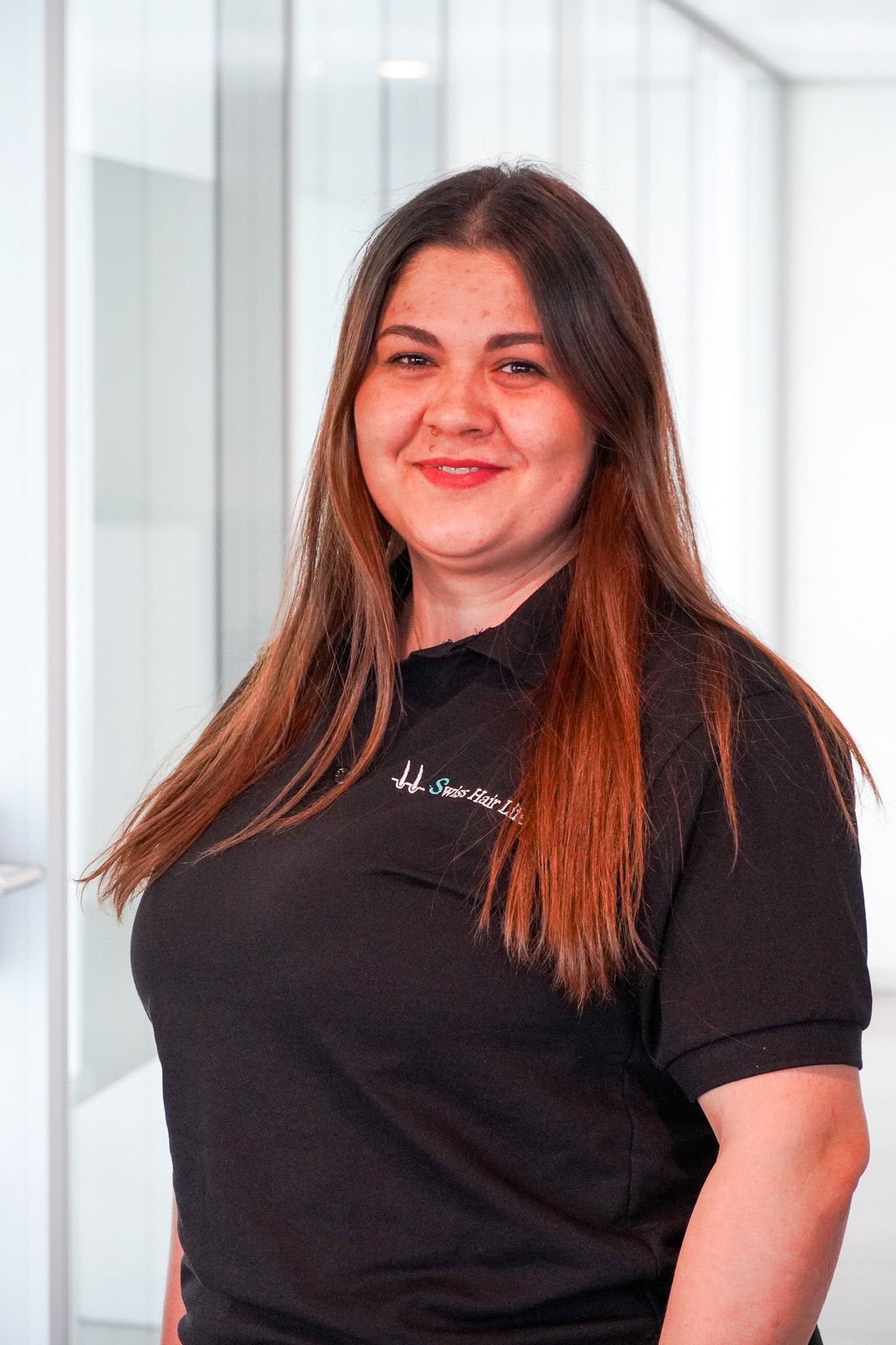Frau mit langen braunen Haaren, lächelnd, in einem schwarzen Poloshirt in einem modernen Büroumfeld.