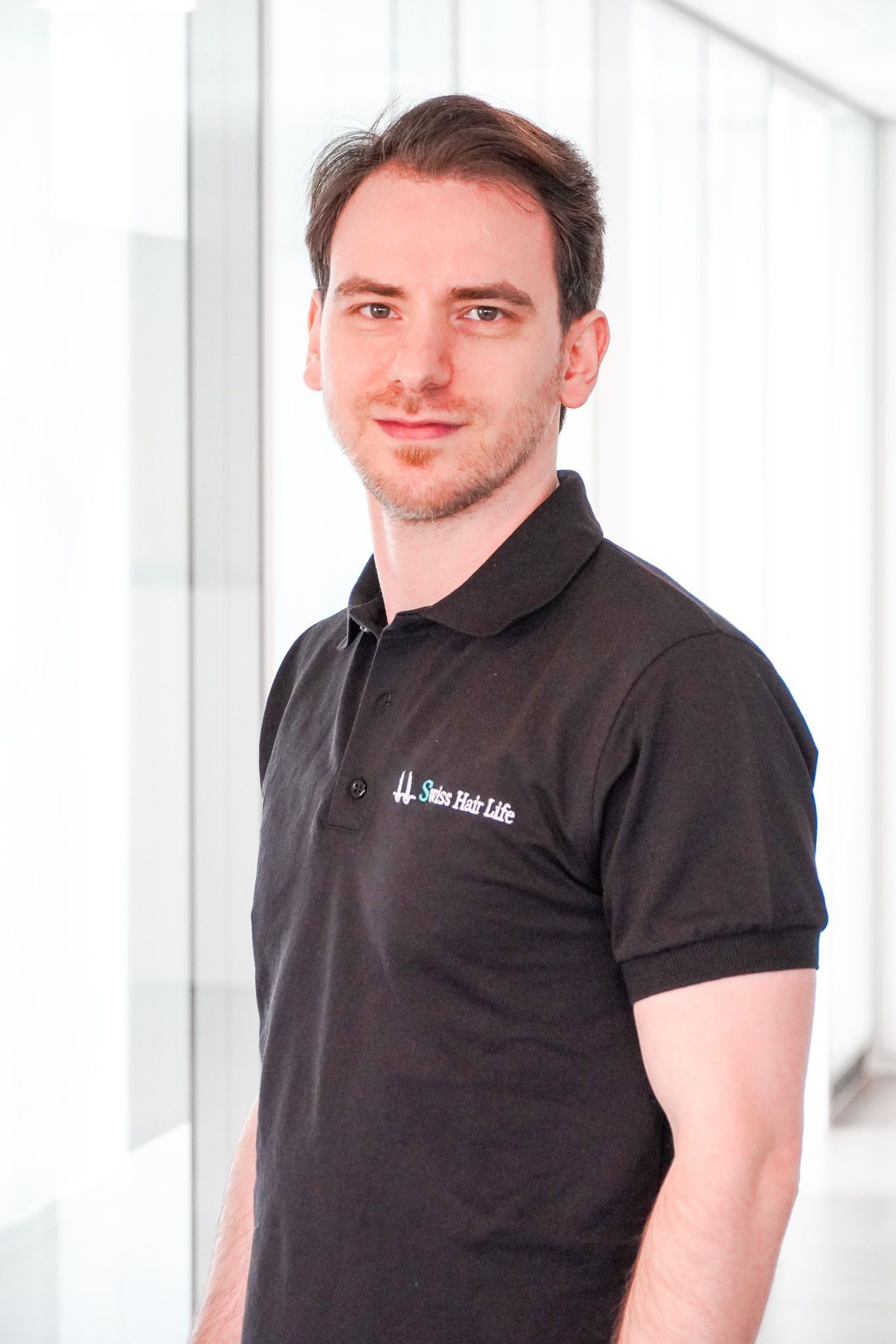 Mann im schwarzen Poloshirt lächelt, steht in einem gut beleuchteten Flur.