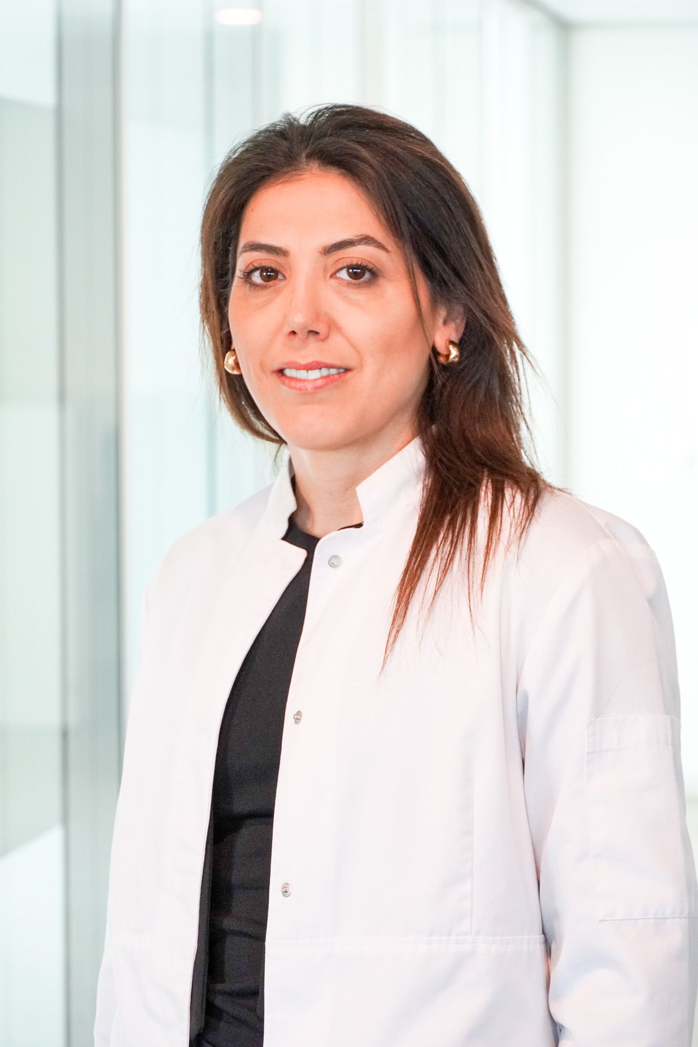 Frau im weißen Laborkittel, lächelnd, steht in einem modernen, hellen Gebäude.