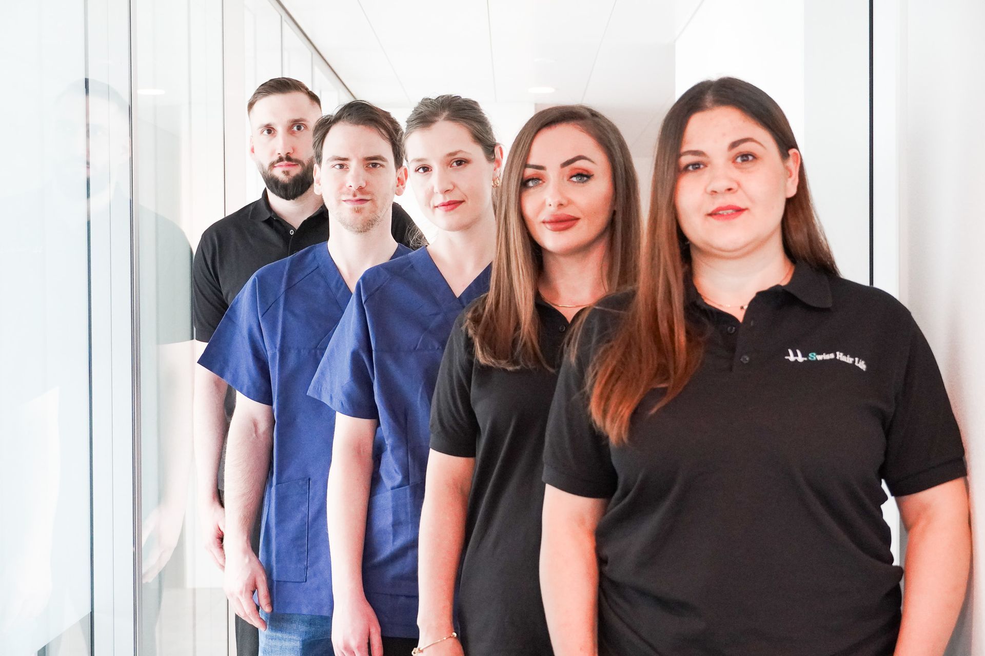 Eine Gruppe von sechs Mitarbeitern des Gesundheitswesens steht in einem Flur und trägt Kittel und Hemden in verschiedenen Farben.