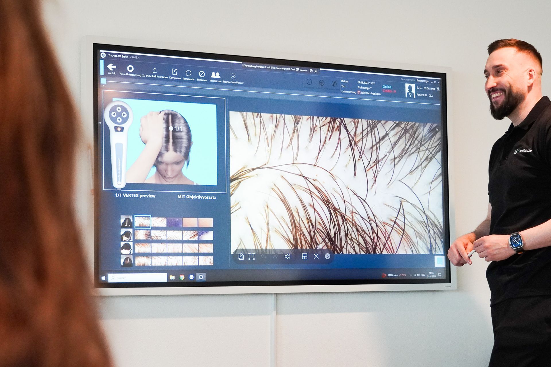 Ein Mann lächelt und zeigt einen Bildschirm mit einer Haaranalyse. Der Bildschirm zeigt eine vergrößerte Ansicht der Haare und der Kopfhaut einer Frau.