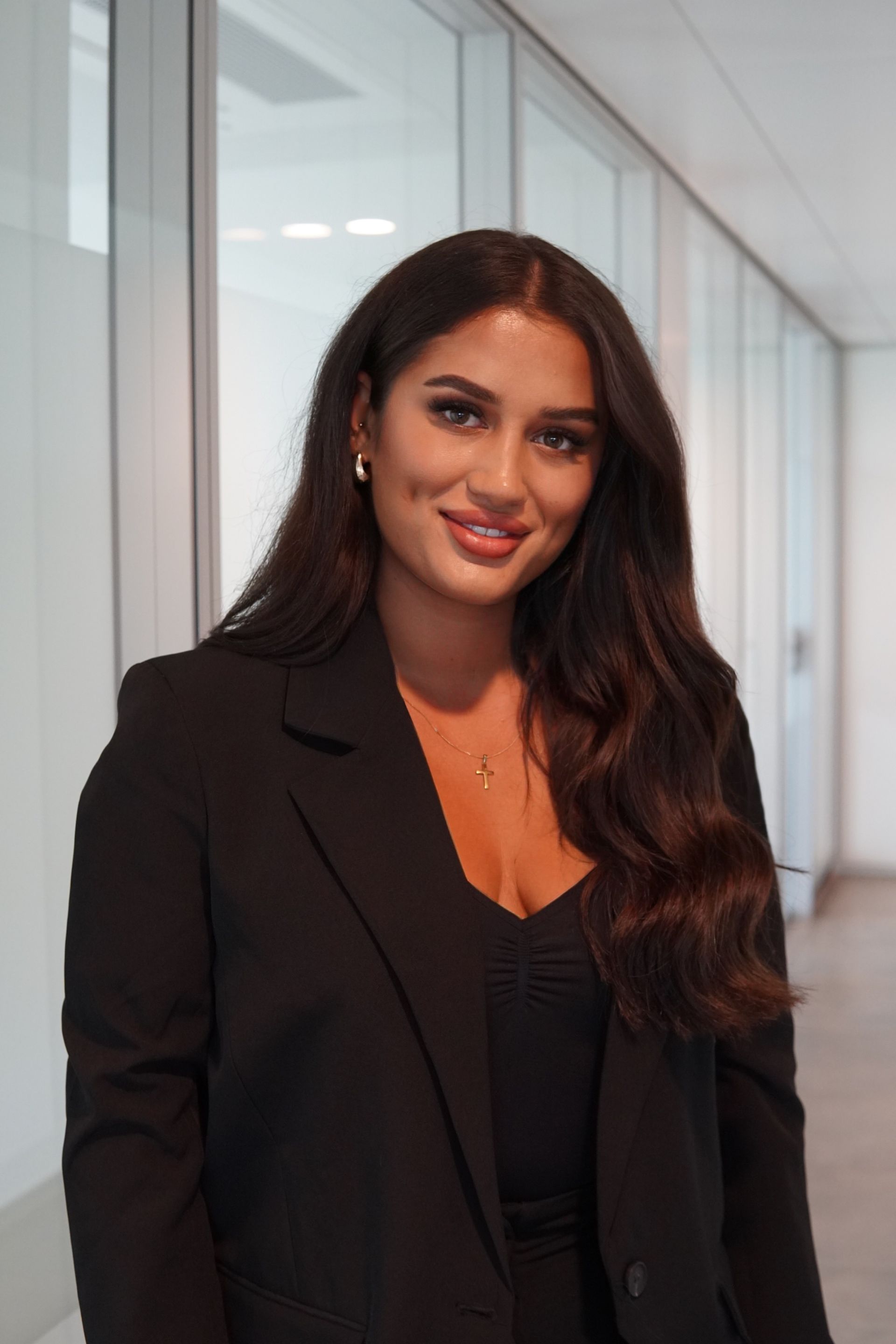 Frau mit langen braunen Haaren, lächelnd, in einem schwarzen Poloshirt in einem modernen Büroumfeld.