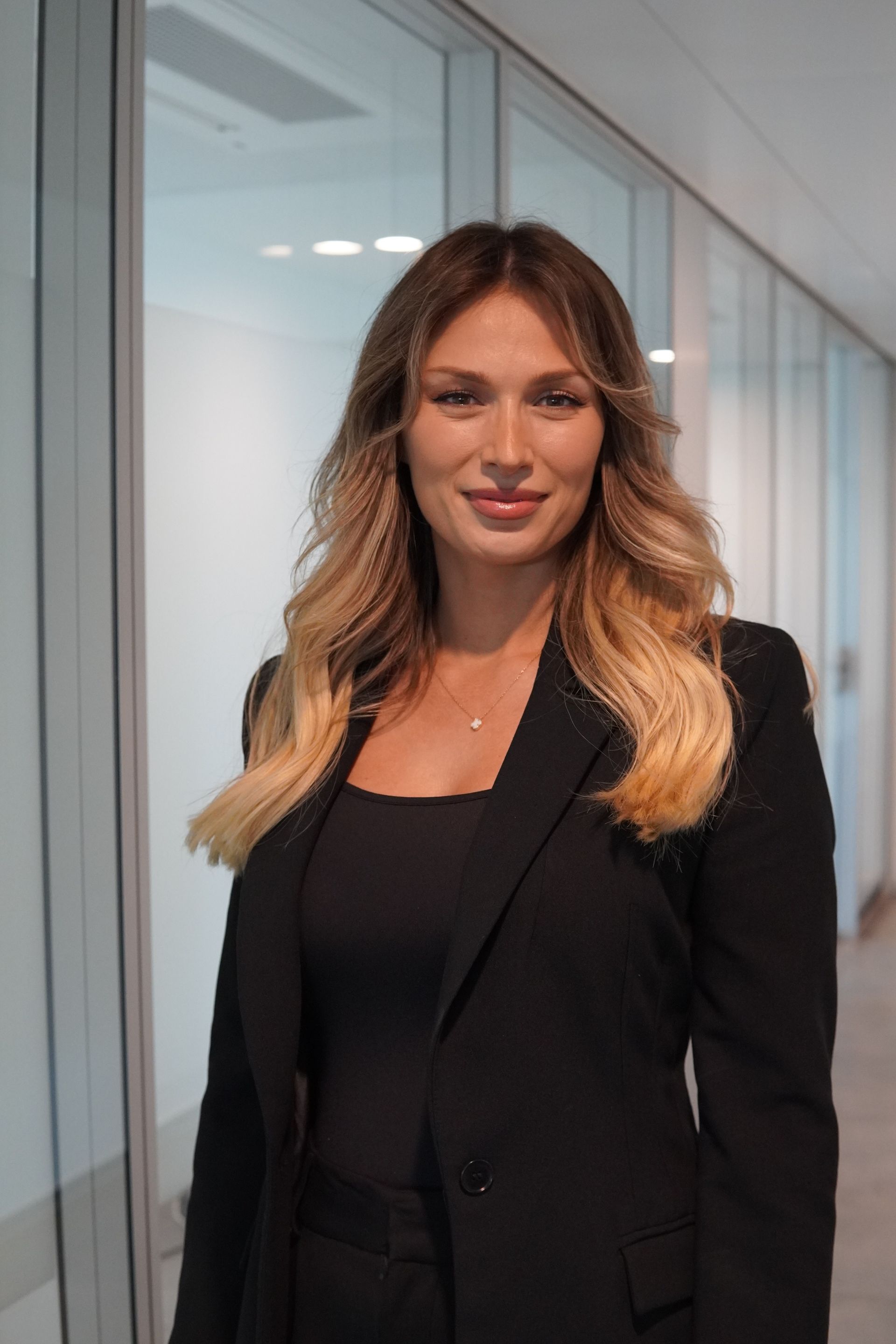 Frau im weißen Laborkittel, lächelnd, steht in einem modernen, hellen Gebäude.