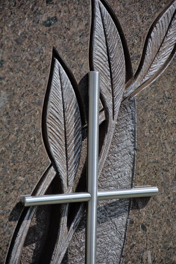 Ein silbernes Kreuz vor eingravierten Blättern auf brauner Granitoberfläche.