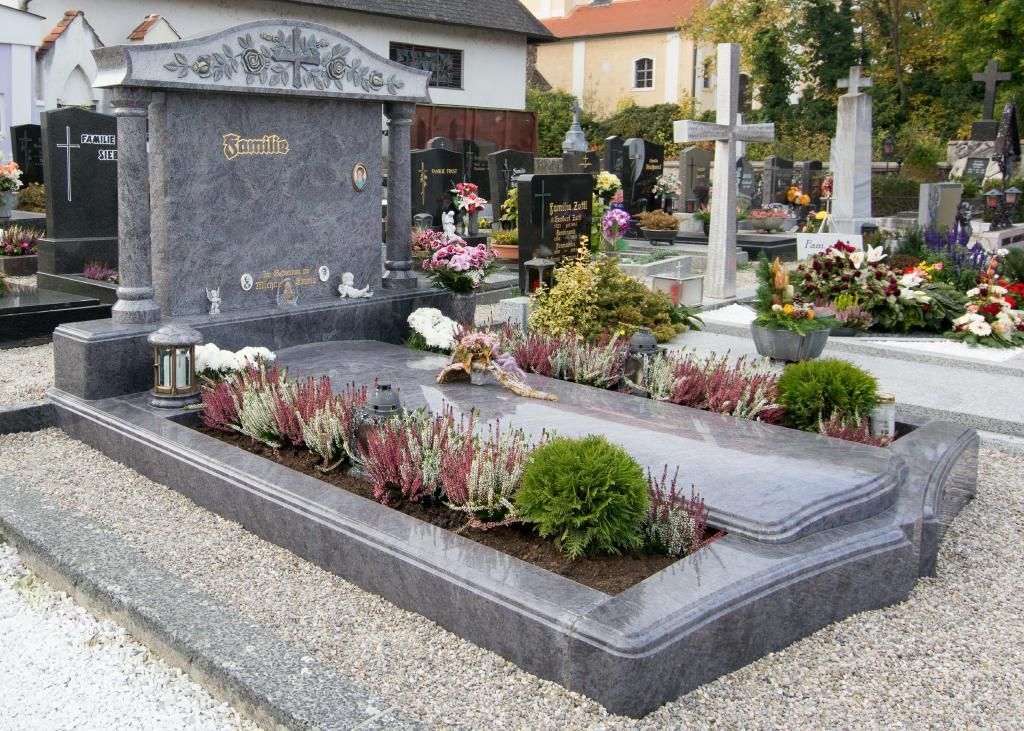 Aufwendig gestalteter, grauer Granitgrabstein mit floralen Verzierungen und Pflanzbeeten auf einem Friedhof.