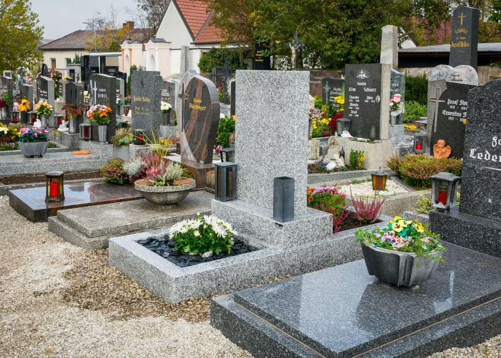 Friedhof mit Granitgrabsteinen und farbenfrohen Blumenarrangements, im Hintergrund ein Gebäude.