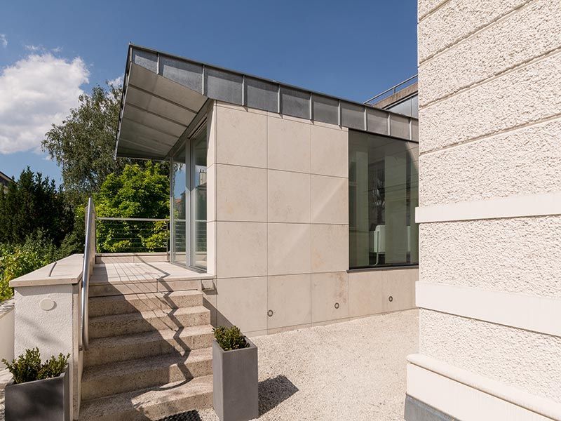 Modernes Gebäude mit Steinfassade, Glasfenstern und Metalldach. Eine Betontreppe führt zum Eingang.