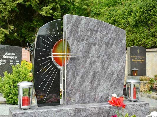 Moderner Granitgrabstein mit stilisiertem Sonnenmotiv, zwei Laternen und roten Blumen.