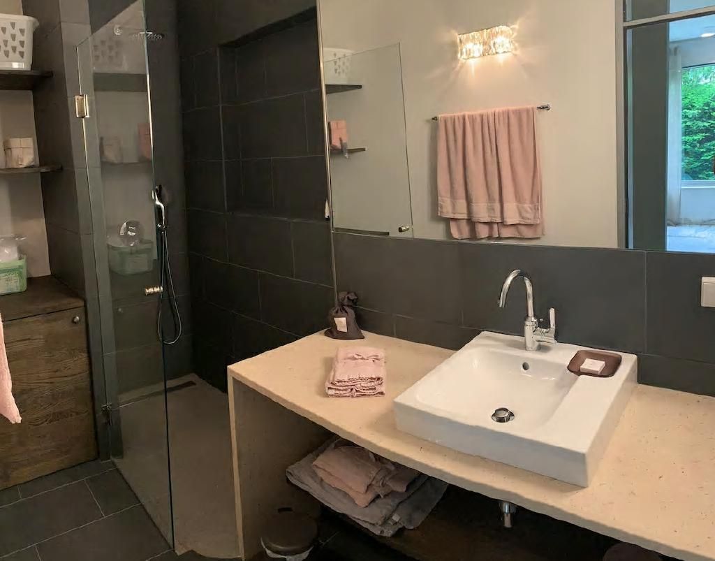 Modernes Badezimmer mit grauen Fliesen, einem großen Spiegel, einem weißen Waschbecken und rosa Handtüchern.