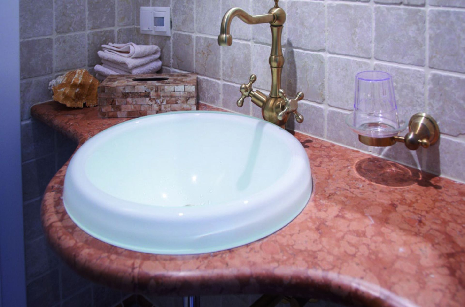 Waschbecken im Badezimmer mit goldenem Wasserhahn auf rötlicher Arbeitsplatte, im Hintergrund kleine Fliesen.