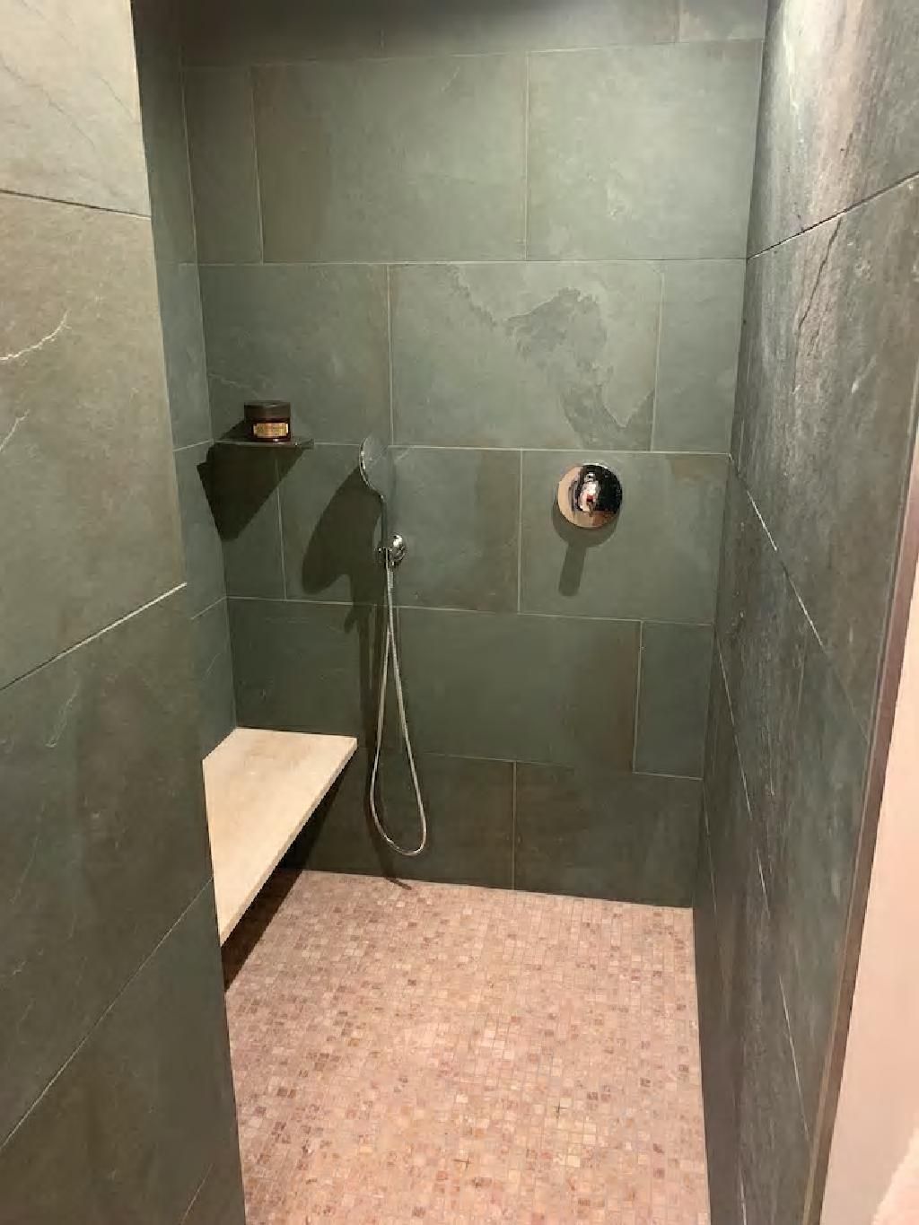 Dusche mit grünen Fliesenwänden, einer Steinbank und Kieselboden. Eine Handbrause hängt daran.