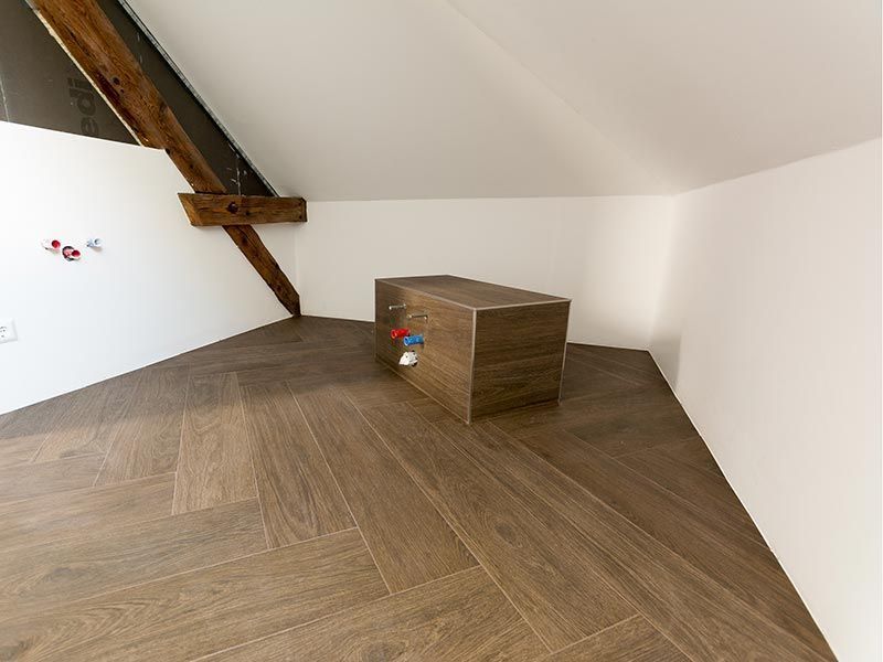 Dachgeschossbadezimmer mit Fliesenboden in Holzoptik, weißen Wänden, freiliegenden Balken und gefliester, kastenartiger Struktur mit Sanitärinstallationen.
